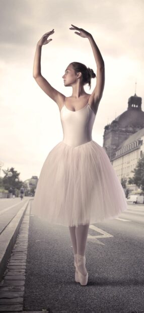 A ballet dancer in a white tutu performs a pose gracefully on a city street during daylight, 2K Phone Wallpaper