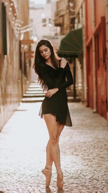 A ballet dancer stands poised on pointe in a black dress on a sunlit street, 4K Mobile Wallpaper