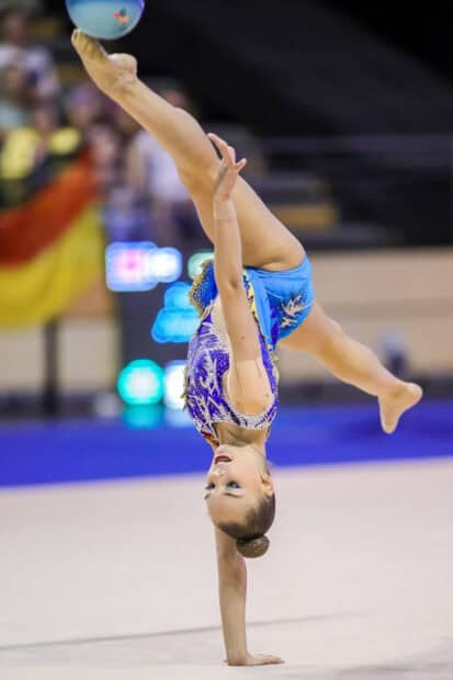 A rhythmic gymnastics athlete performs a one handed handstand with a ball in midair, HD Mobile Wallpaper