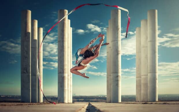 A rhythmic gymnast leaps in front of stone columns holding a red and white ribbon, HD Desktop Wallpaper