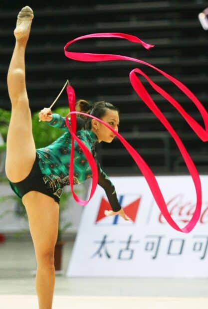 A rhythmic gymnast performs a high leg lift while twirling a red ribbon in competition, HD Mobile Wallpaper