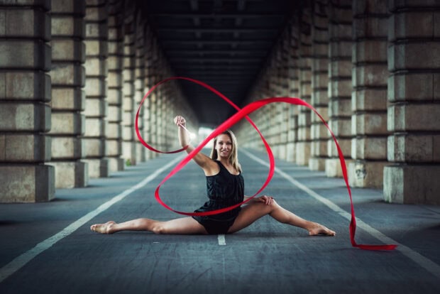 A rhythmic gymnastics athlete performs a split with a flowing red ribbon under a bridge, HD Desktop Wallpaper