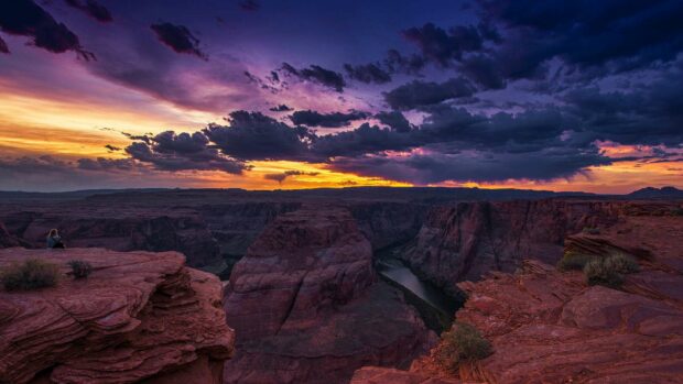 A person is sitting on red rocks overlooking the Grand Canyon under a colorful sunset sky, HD Desktop Wallpaper