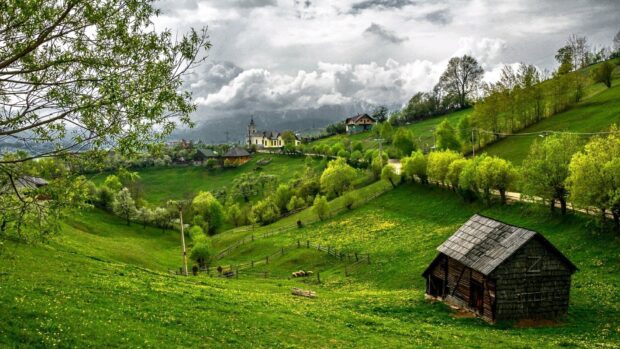 A village is surrounded by lush green hills and blooming trees under a cloudy sky, HD Desktop Wallpaper
