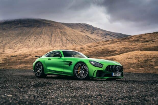 A green Mercedes Benz AMG GT is parked on a gravel road with mountains in the background under a cloudy sky, 2K Desktop Wallpaper