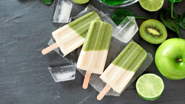 Three green and white popsicles on a dark surface surrounded by kiwi, lime, and ice cubes, 4K Desktop Wallpaper