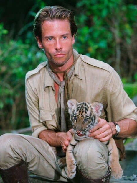 Guy Pearce holding a baby tiger in a jungle setting looking serious, 2K Desktop Wallpaper