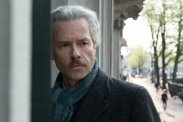 A man with gray hair and a mustache stands focused on the street, wearing a patterned scarf and dark coat