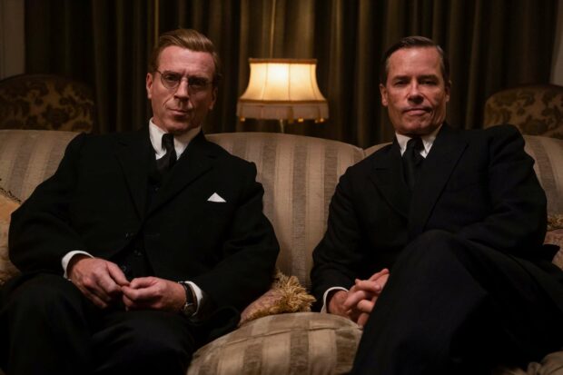 Guy Pearce sitting with a man on a couch wearing black suits under warm lighting