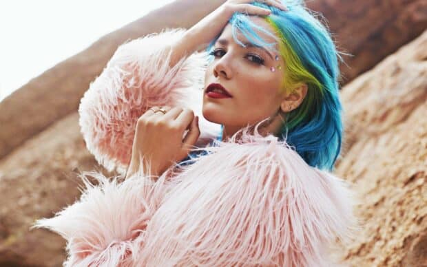 Halsey with blue and yellow hair wearing a pink fur coat against a rocky background, 2K Desktop Wallpaper