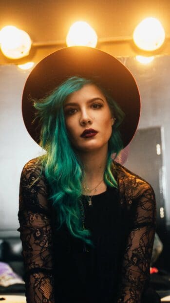 A woman with teal hair wearing a black hat and lace top under warm lighting, 2K Mobile Wallpaper