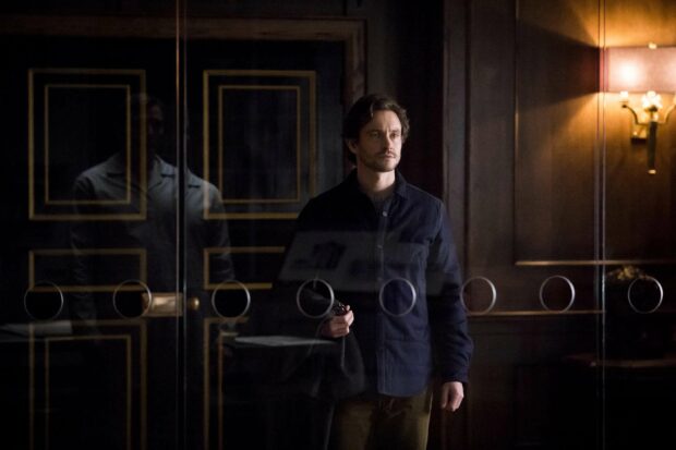 Mads Mikkelsen stands thoughtfully in a dimly lit room with reflections on glass, 2K Desktop Wallpaper