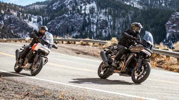 Two riders on Harley Davidson Pan America 1250 motorcycles are turning on a mountain road with snowy trees in the b