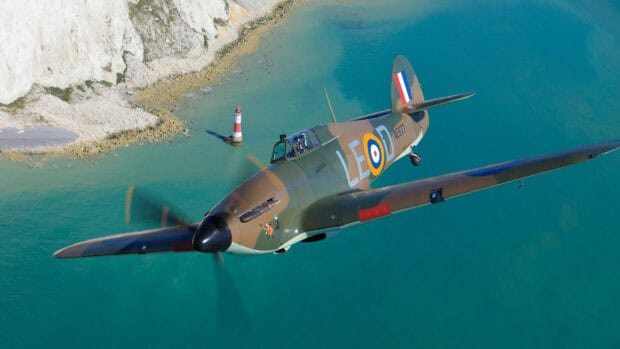 A Hawker Hurricane aircraft flies above blue waters near a rocky shore with a lighthouse, HD Desktop Wallpaper