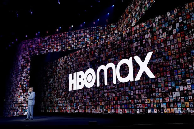 A speaker presents in front of a large screen displaying HBO Max with multiple show thumbnails, HD Desktop Wallpaper