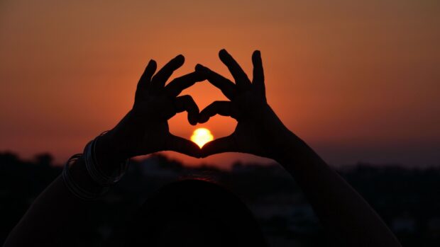 A person forms a heart shape with their hands framing the sun at sunset, 4K Desktop Wallpaper
