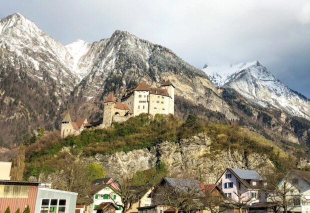 A historic castle stands on a hill surrounded by houses and snowy mountains in Liechtenstein wallpapers, 2K Desktop Wallpaper