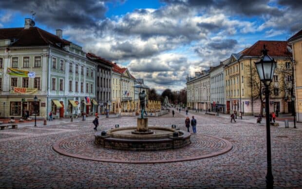 A historic town square in Estonia shows people walking among old buildings under a dramatic cloudy sky, 2K Desktop Wallpaper