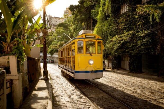 A yellow tram travels through a sunlit street in Rio De Janerio with lush greenery nearby, 2K Desktop Wallpaper