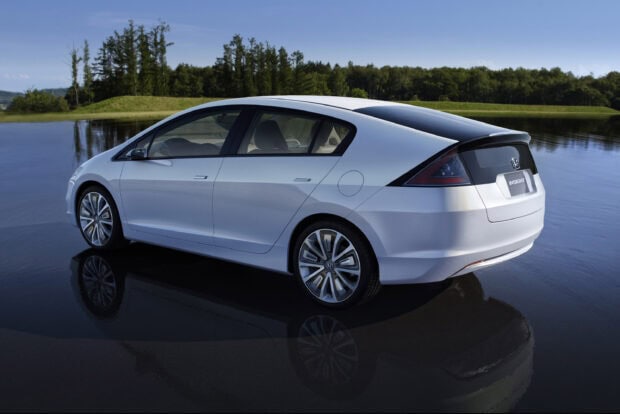 A white Honda Insight hybrid car is parked near a calm lake reflecting the vehicle and surrounding trees, 2K Desktop Wallpaper