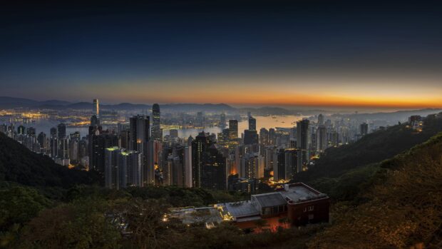 A panoramic view of the Hong Kong skyline at dusk showing illuminated buildings and distant mountains, 4K Desktop Wallpaper