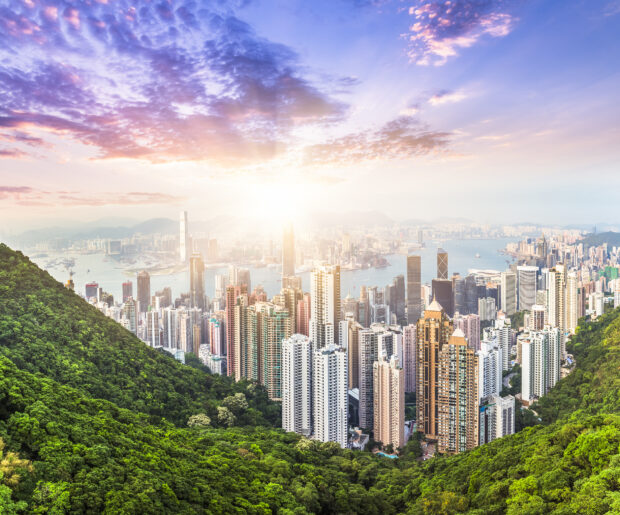 The Hong Kong skyline features towering skyscrapers and lush green hills under a colorful sunrise sky, HD Desktop Wallpaper