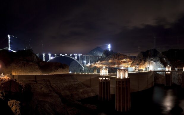 The Hoover Dam stands illuminated at night with the surrounding rocky landscape and a dark cloudy sky, HD Desktop Wallpaper