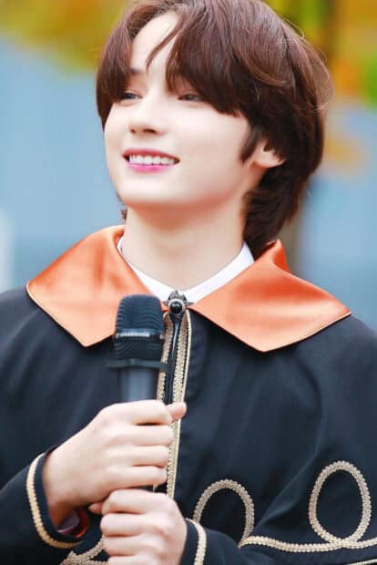 Huening Kai is smiling warmly while holding a microphone and wearing a decorative outfit, HD Mobile Wallpaper