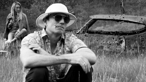 A man wearing a hat and sunglasses with a cigar near a damaged car in a grassy field, HD Desktop Wallpaper