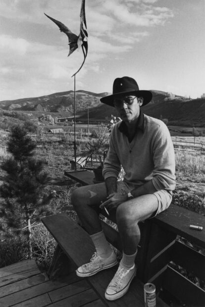 A man wearing sunglasses and a hat sits outdoors with mountains in the background, 2K Desktop Wallpaper