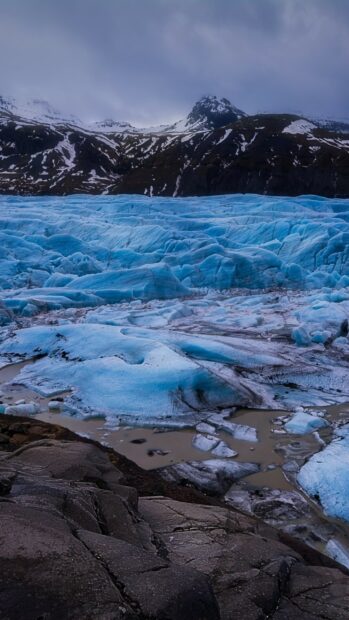 A vast glacier landscape showcases blue ice formations with snowy mountains in the background, 2K Mobile Wallpaper