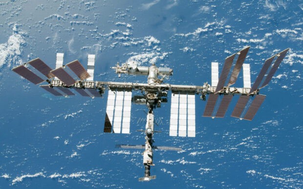 The space station is orbiting Earth with visible solar panels and cloud patterns below, 2K Desktop Wallpaper