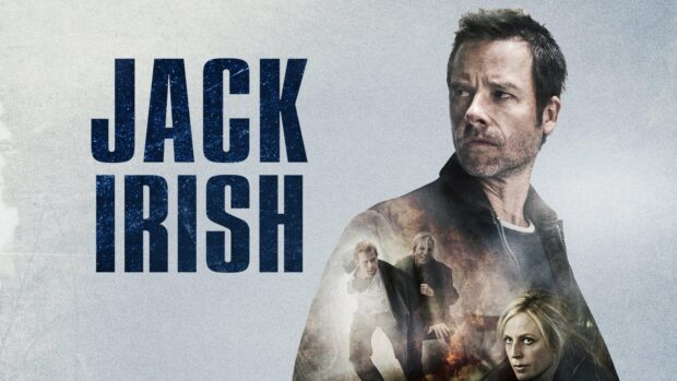 A man with a serious expression stands with other characters appearing in a smoky background from the Jack Irish series
