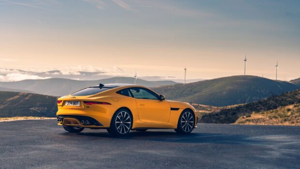 A yellow Jaguar F Type is parked on a mountain road with wind turbines in the background, 2K Desktop Wallpaper