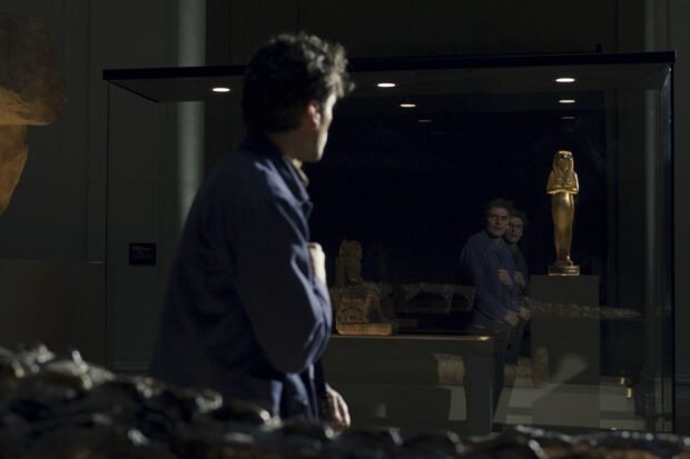 Jake Lockley watches his reflection beside a golden statue inside a museum display case, HD Desktop Wallpaper