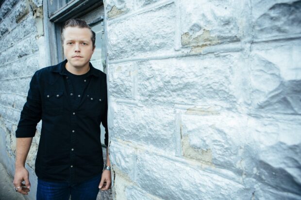 Jason Isbell is standing next to a textured stone wall wearing a black shirt, HD Desktop Wallpaper