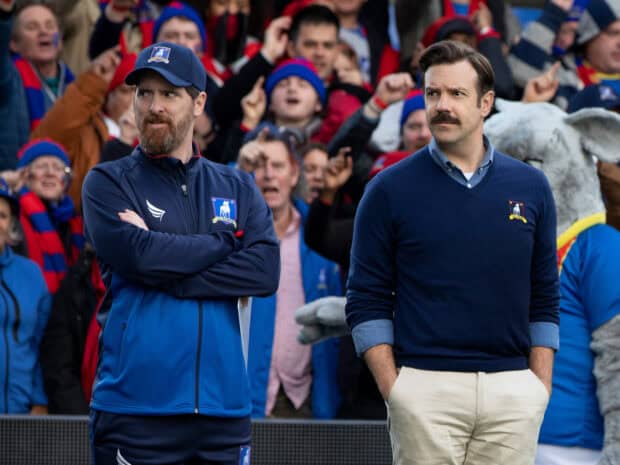 Jason Sudeikis watches a football match with a man in a blue jacket during a crowded event, 2K Desktop Wallpaper