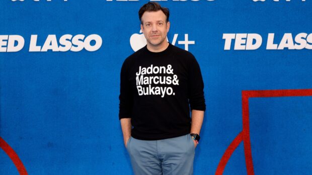 Jason Sudeikis Wallpapers in a black sweatshirt standing against a blue Ted Lasso backdrop, HD Desktop Wallpaper