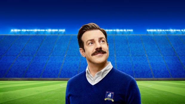 Jason Sudeikis is standing on a green field with bright lights and blue seats behind him, HD Desktop Wallpaper