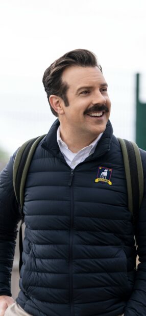 Jason Sudeikis is smiling while wearing a navy blue jacket and carrying a backpack, 2K Phone Wallpaper