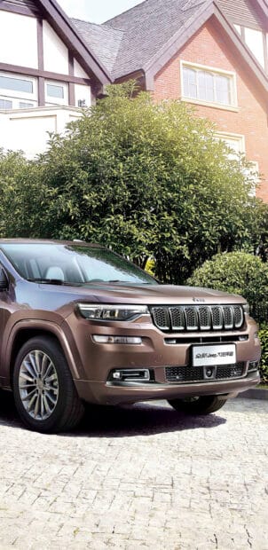 A brown Jeep Commander is parked on a driveway near green bushes and a house, 2K Phone Wallpaper