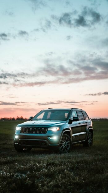 A Jeep Grand Cherokee drives through a field under a colorful sunset sky with headlights on, HD Phone Wallpaper