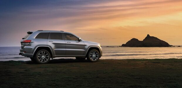 A silver Jeep Grand Cherokee is parked near the ocean with a scenic island and sunset sky in the background, 2K Desktop Wallpaper
