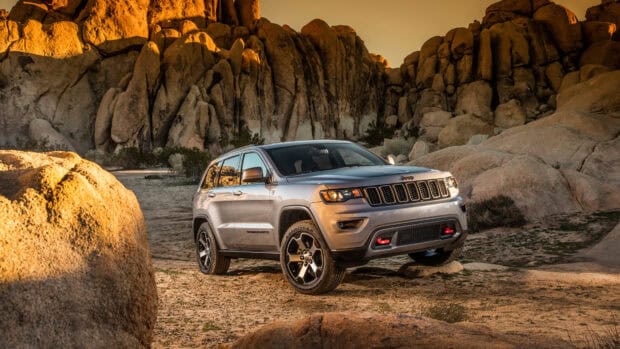A silver Jeep Grand Cherokee is parked on rough desert terrain with large rocky formations in the background, 4K Desktop Wallpaper