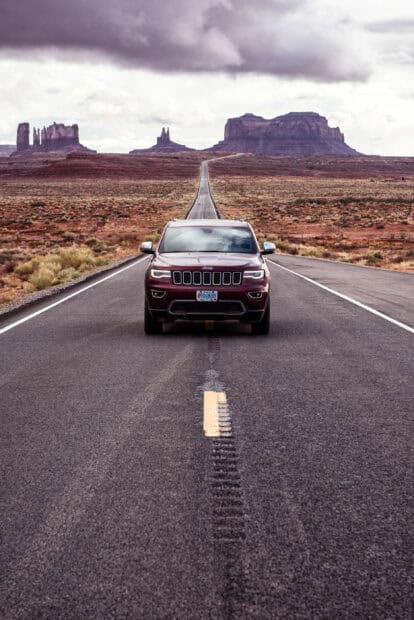 A Jeep Grand Cherokee drives down a long desert highway with rocky formations in the background, 2K Phone Wallpaper