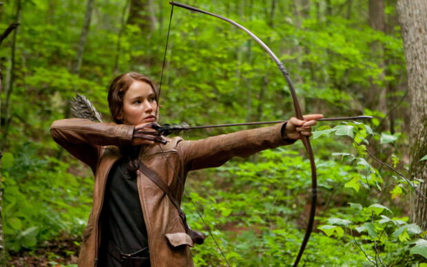 Jennifer Lawrence aiming a bow and arrow in a forest scene from The Hunger Games, HD Desktop Wallpaper