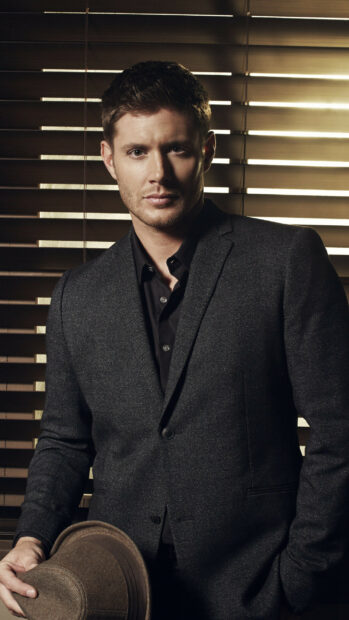 Jensen Ackles in a dark suit holding a brown hat in front of window blinds, 2K Mobile Wallpaper