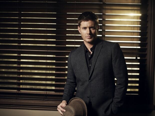 Jensen Ackles stands confidently in a dark suit holding a hat by the window blinds, HD Desktop Wallpaper
