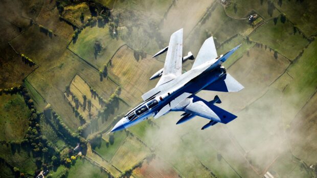 A jet fighter is flying high over green farmland below on a clear day, 2K Desktop Wallpaper