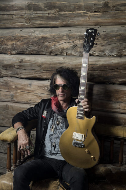 A man wearing sunglasses holds a Gibson guitar while sitting against a wooden wall for Joe Perry Wallpapers, 2K Desktop Wallpaper
